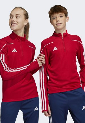 Rode sportjacks met halve rits, voorzien van witte strepen op de mouwen en een Adidas-logo. Gecombineerd met blauwe sportbroeken.