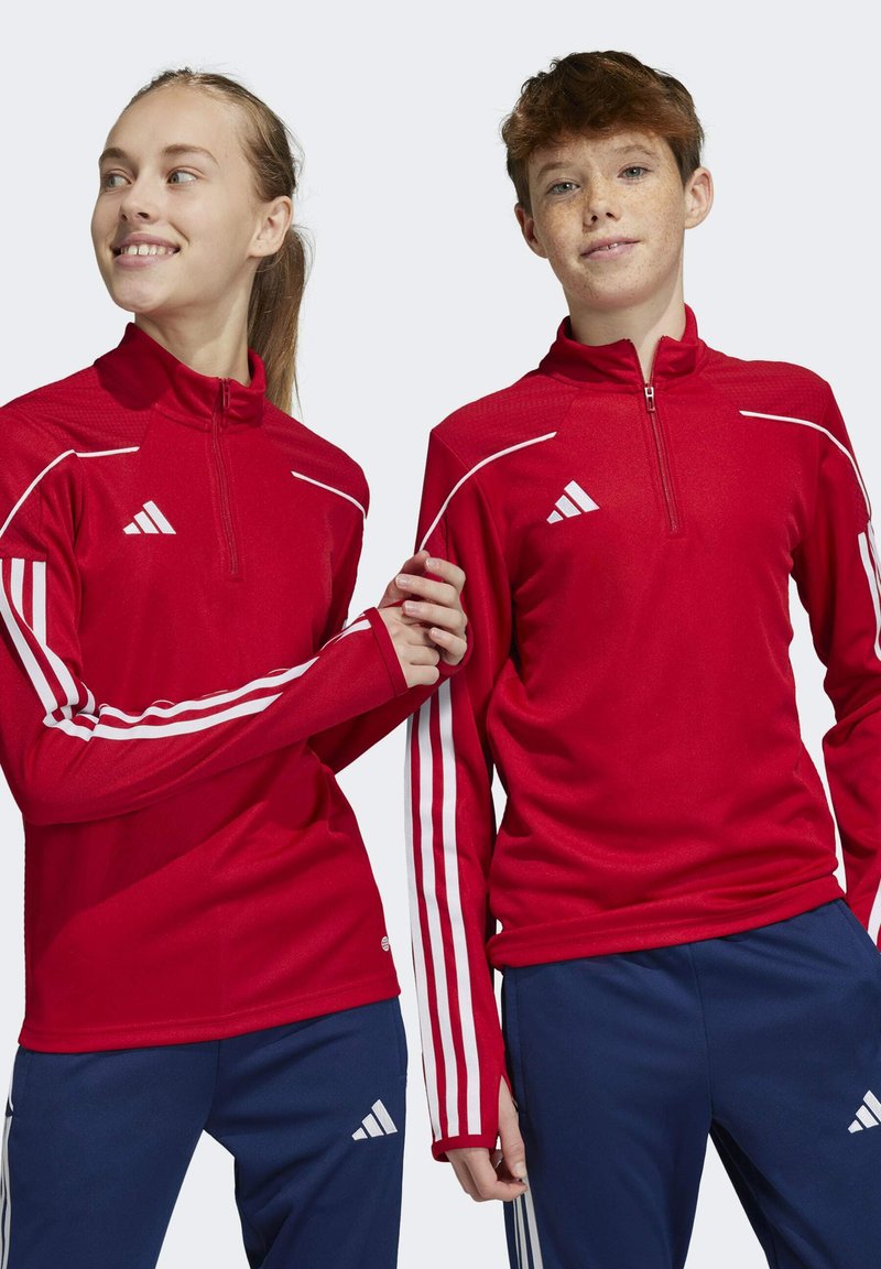 Vestes de sport rouges avec demi-zip, comportant des rayures blanches sur les manches et un logo Adidas. Associées à un pantalon de sport bleu.