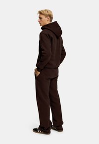 Brun hettegenser og matchende joggebukser i myk stoff, med stor hette, avslappet passform og ribbede mansjetter. Skoene har tre hvite striper.