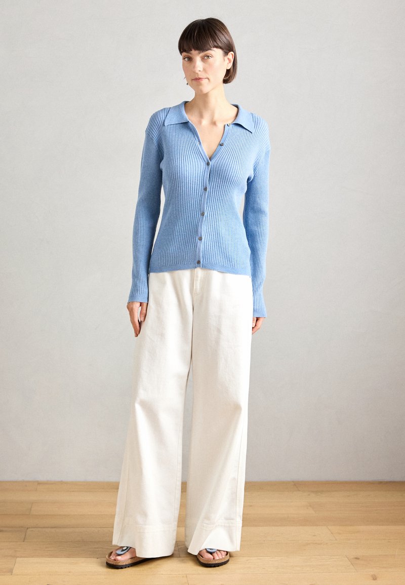 Hellblau gestrickte Cardigan mit Knopfverschluss und spitzem Kragen, kombiniert mit weißen, weit geschnittenen Hosen und schwarzen Sandalen. Glatte Textur.