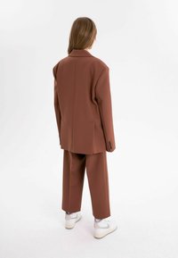 Brun oversized kavaj och matchande byxor i strukturerad tyg. Vita sneakers med grå detaljer kompletterar outfiten.