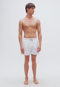 Λευκά μαγιό σορτς από ελαφρύ ύφασμα με ελαστική μέση και ρυθμιζόμενο σκοινί. Το μοντέλο στέκεται ξυπόλητο, κοιτάζοντας προς τα αριστερά.