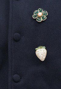 Två broscher nålade på ett mörkt tyg: den ena är en grön blomma med en pärlcentral, den andra en vit jordgubbe med gröna accenter och gyllene detaljer.