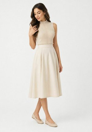 Woman with long dark hair wearing a sleeveless beige ribbed top, cream midi skirt, and beige flat shoes, standing against a plain background.