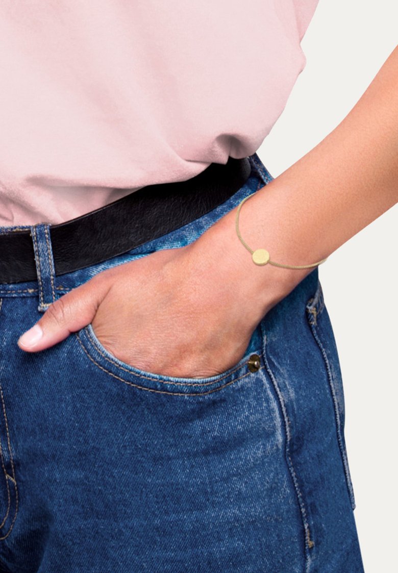 Guldtonad armband med en cirkulär berlock, bärs på en handled, ihop med en ljusrosa skjorta och blå denimjeans, med en framficka.
