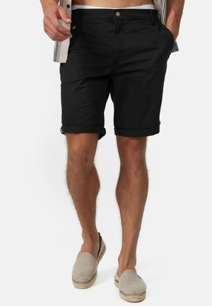 Mand iført sorte shorts med opviklede kanter, beige espadrillos og en delvist synlig ternet skjorte med hænderne i lommerne.