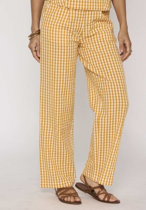 Donna con pantaloni ampi a quadretti gialli e bianchi e sandali bassi marroni, con bracciali e anelli alle mani.
