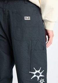 Pantaloni de lucru negri, cu un buzunar posterior, design grafic în formă de explozie albă pe coapsă și detalii de cusătură în contrast.