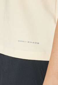 Jasnożółta koszula OMNI-SHADE o miękkiej teksturze, z szarym logo blisko dołu, w połączeniu z ciemnym materiałem poniżej.