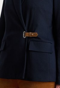 Blazer blu navy con chiusura frontale a fibbia in pelle marrone e tasche a patta, indossato su pantaloni in velluto a coste marrone.