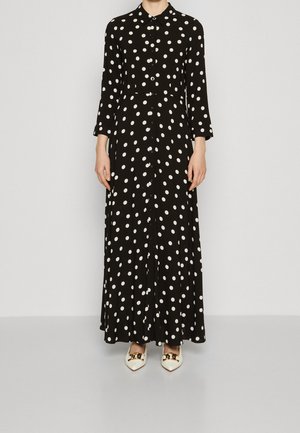 Zwarte maxi-jurk met lange mouwen en witte polka dots, met knopen aan de voorkant en een kraag, gedragen met witte puntige schoenen met gouden kettinkdetail.