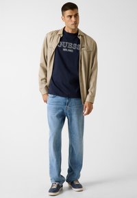 Beiges Langarmshirt über einem marineblauen T-Shirt mit "GUESS"-Logo, kombiniert mit hellblauen Jeans und marineblauen Sneakers mit braunen Akzenten.