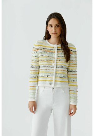 Femme aux longs cheveux bruns portant un cardigan tricoté à rayures blanches, jaunes et oranges ainsi qu’un pantalon blanc, debout devant un fond uni.