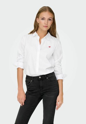 Chemise blanche à manches longues avec un petit logo en forme de cœur rouge sur la poitrine gauche. Associée à un jean noir taille haute.