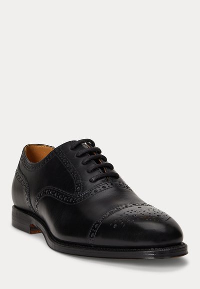 Zapato de vestir de hombre de cuero negro con detalles tipo brogue, cordones cerrados y un tacón bajo apilado sobre un fondo blanco.