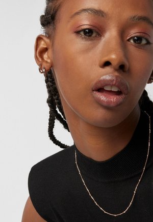 Nahaufnahme einer Frau mit geflochtenem Haar, die kleine Creolen, eine dünne Kettenhalskette und ein schwarzes ärmelloses Oberteil mit hohem Ausschnitt trägt.
