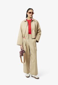 Giacca beige cropped con dettagli rossi e verdi, abbinata a una maglietta rossa e pantaloni beige a gamba larga. Gli accessori includono una piccola borsa bordeaux e occhiali da sole.
