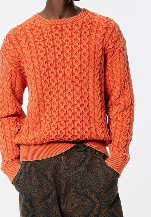 Personne portant un pull en maille torsadée orange vif et un pantalon à motif cachemire vert foncé, les mains dans les poches.