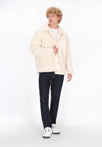 Cremfarbener, strukturierter Fleece-Jacke mit großem Kragen, vorderen Knöpfen und einer Brusttasche, kombiniert mit dunkler Jeans und weißen Turnschuhen.
