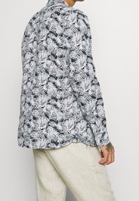 Uomo che indossa una camicia a maniche lunghe con motivo foglia tropicale in bianco e nero e pantaloni beige chiari, con una mano tatuata visibile.