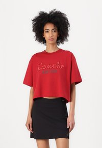 Rotes, kurzes T-Shirt mit kurzen Ärmeln, das ein erhabenes Logo "Coach New York" in Schwarz und Leopardenmuster trägt. Kombiniert mit einem schwarzen Rock.