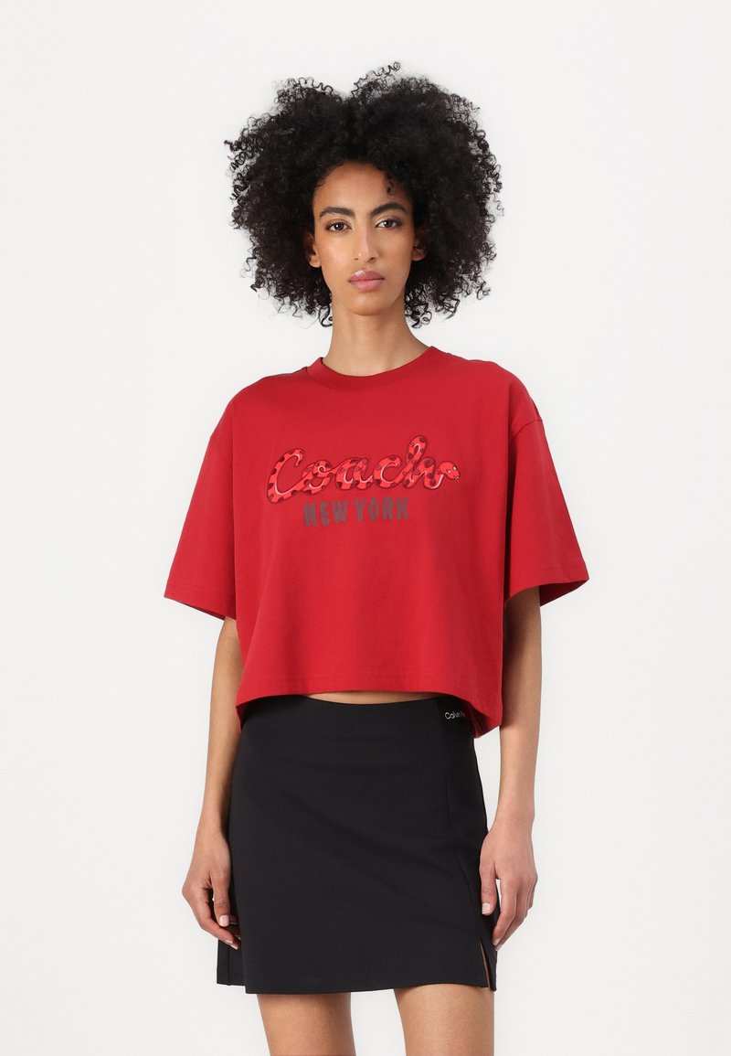 Rotes, kurzes T-Shirt mit kurzen Ärmeln, das ein erhabenes Logo "Coach New York" in Schwarz und Leopardenmuster trägt. Kombiniert mit einem schwarzen Rock.