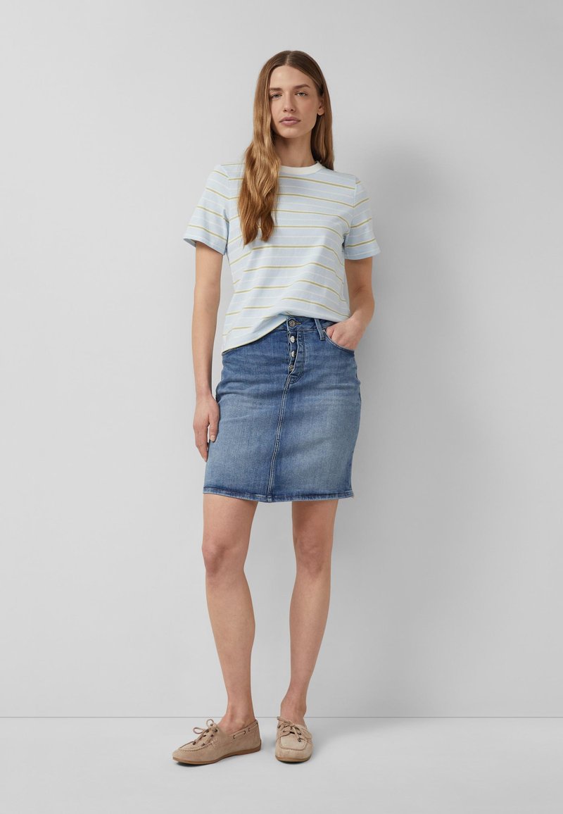 Femme debout, main dans la poche, portant une chemise à manches courtes bleu clair rayée, une jupe en jean boutonnée et des chaussures décontractées beige.