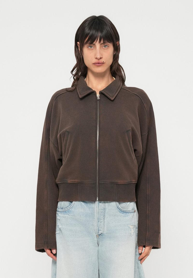 Veste courte marron en tissu doux, équipée d'une fermeture éclair, d'un col pointu et de poches latérales, avec des détails de coutures apparentes.