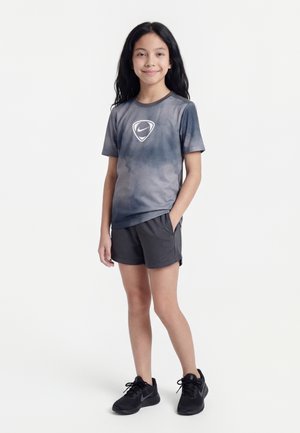 Young girl with long dark hair wears a gray Nike sports shirt, black shorts, and black Nike sneakers, standing with one hand in pocket.