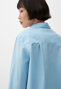 Lichtblauw, langwerpig shirt met een kraag, met een ontspannen pasvorm en zachte, textuurrijke stof. Een rugplooidetail voegt volume toe.