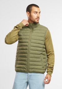 Grüne gesteppte Weste mit hohem Kragen und Reißverschluss, über einem olivfarbenen Langarmpullover getragen. Lässige Passform, strukturierter Stoff, strukturiertes Design.