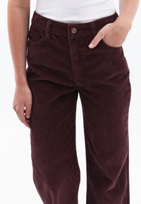 Pantalon en velours côtelé bordeaux avec un motif texturé et strié, taille haute, design à cinq poches et fermeture à bouton avec logo.