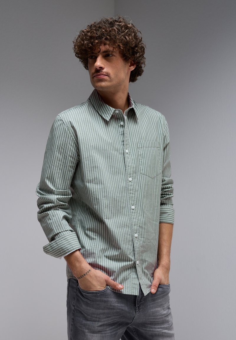 Vihreä ja valkoinen raidallinen nappikauluspaita, jossa on yksi rintatasku ja ylöskäännetyt hihat, yhdistettynä tummanharmaisiin farkkuihin.