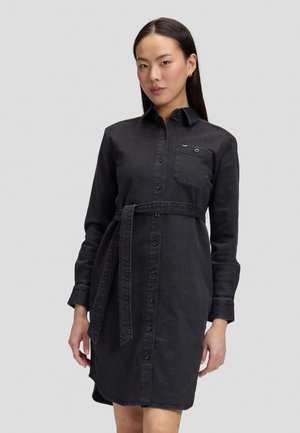 Vrouw met lang donker haar die een zwarte denim overhemdjurk draagt met knopen, een riem en een borstzak, staand tegen een effen grijze achtergrond.