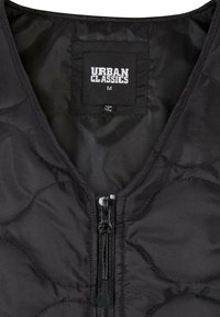 Veste matelassée noire avec fermeture éclair à l'avant et étiquette de marque Urban Classics indiquant la taille medium à l'intérieur du col.