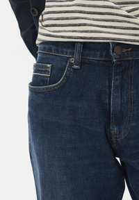 Temno modri kavbojki imajo klasičen dizajn s petimi žepi in kontrastnim šivanjem. Tekstura je gladka, z nekoliko sijočim videzom.