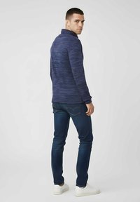 Jersey de textura en azul marino con cuello alto, combinado con vaqueros azul oscuro y zapatillas blancas. Vista desde atrás, mostrando el ajuste y el diseño.