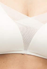 Λευκό σουτιέν με απαλές μπανέλες και διαφανές δίχτυ σε σταυρωτό σχέδιο στο κέντρο σε γυμνό στήθος.