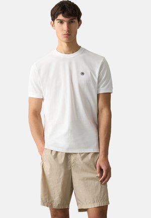 T-shirt bianca a maniche corte con un piccolo logo circolare sul petto, abbinata a pantaloncini beige chiaro realizzati in tessuto strutturato.