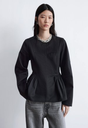 Black long-sleeve top with a fitted bodice and peplum detail, made from a smooth cotton blend. Paired with gray denim pants and layered jewelry.