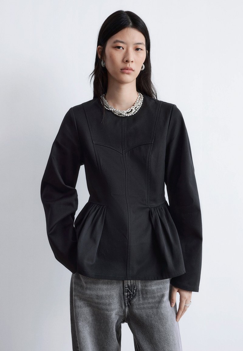 Schwarzes Langarmoberteil mit figurbetontem Oberteil und Volant-Detail, aus einem glatten Baumwollgemisch. Kombiniert mit grauer Jeanshose und geschichteten Schmuckstücken.