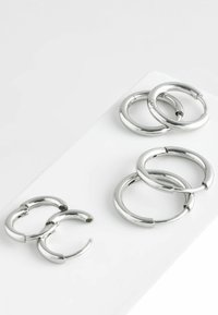 Cercei din metal argintiu în două dimensiuni. Cerceii mai mici sunt grupați împreună, în timp ce cei mai mari sunt aranjați separat. Finisaj neted și strălucitor.