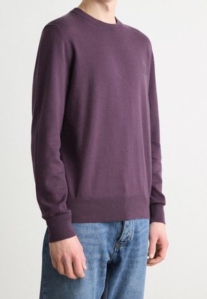 Homme portant un pull violet uni à col rond et un jean bleu, debout devant un fond clair, les bras détendus le long du corps.