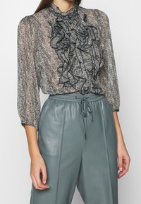 Blusa translúcida con frente acampanado; patrón salpicado en beige y azul marino. Combinada con pantalones de cuero de talle alto en verde azulado claro, que presentan una cinturilla con cordón.