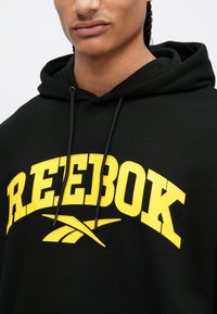 Čierna mikina s kapucňou so veľkým žltým logom "REEBOK" a grafickým dizajnom. Má kapucňu so šnúrkou a mäkkú textúrovanú tkaninu. Detailný pohľad.