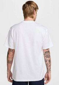 Camiseta blanca de algodón con mangas cortas, ajuste relajado, cuello redondo y una textura suave. Tatuajes visibles en los brazos.