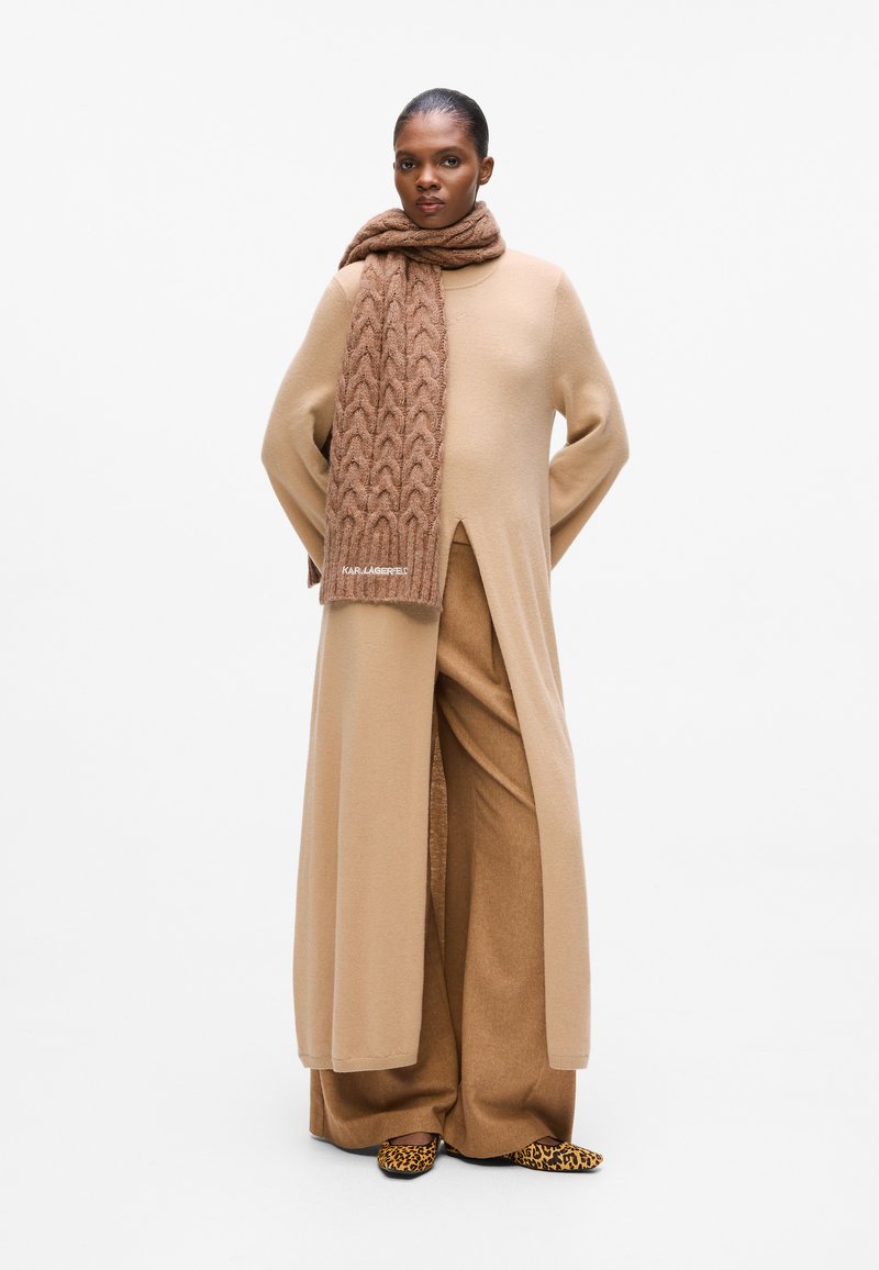 Manteau beige long avec une fente profonde à l'avant, assorti à un pantalon large. Écharpe en tricot marron avec un motif texturé et chaussures à imprimé animal.
