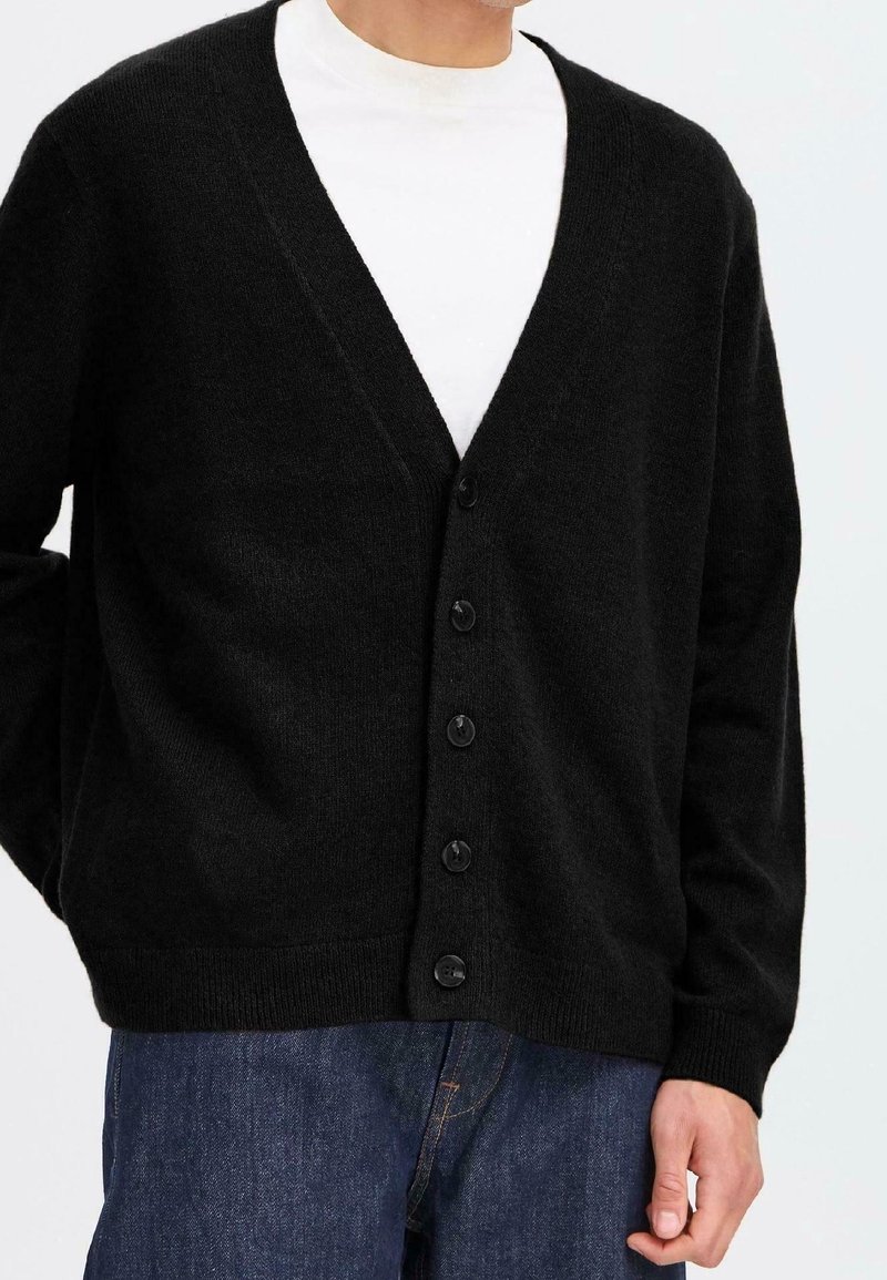 Homme portant un cardigan noir boutonné sur un T-shirt blanc et un jean bleu, debout avec une main détendue sur le côté.