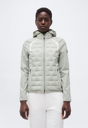 Personne portant une veste matelassée à capuche gris clair avec des poches zippées et un pantalon blanc, face à l'avant sur un fond uni.