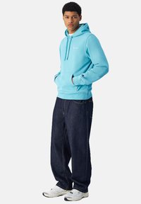 Sudadera azul clara con bolsillo tipo canguro y capucha con cordón, combinada con jeans de pierna ancha azul oscura y zapatillas deportivas blancas.